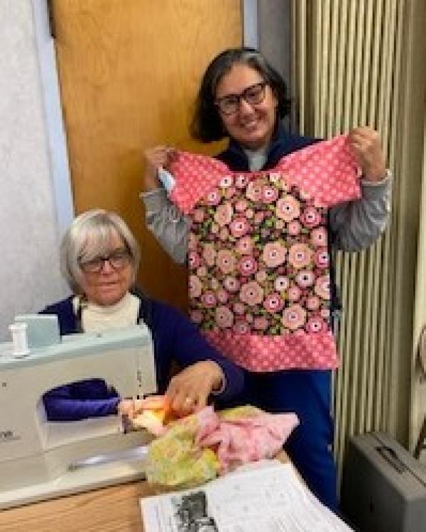 Joan Feffer continues to sew while Merriam Rogers shows off one of the completed dresses.